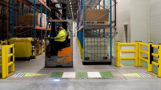 Forklift truck in a warehouse driving towards a zebra crossing and 2 automated projected safety signs warning pedestrians.